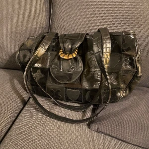 Y2k vintage väska - Säljer en cool svart axelväska med unikt mönster och detaljer. Väskan har en dekorativ knäppning framtill och långa axelremmar. Får plats med A4 så perfekt till skolan!