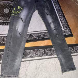 Säljer ett par grå jeans från Philipp Plein. De har en cool distressed look med slitningar och en unik grön detalj på ena benet. Jeansen har en knappgylf och är i en straight stil. Perfekta för en avslappnad men ändå edgy look.