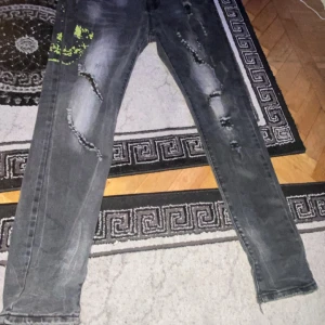 Grå jeans från Philipp Plein - Säljer ett par grå jeans från Philipp Plein. De har en cool distressed look med slitningar och en unik grön detalj på ena benet. Jeansen har en knappgylf och är i en straight stil. Perfekta för en avslappnad men ändå edgy look.