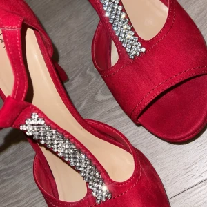 Röda klackskor med strassdetaljer - Snygga röda klackskor från Catwalk i mocka med glittrande strassdetaljer på remmen. Perfekta för festliga tillfällen! De har en bekväm klack och en öppen tå-design som ger en elegant look. Använt en gång 