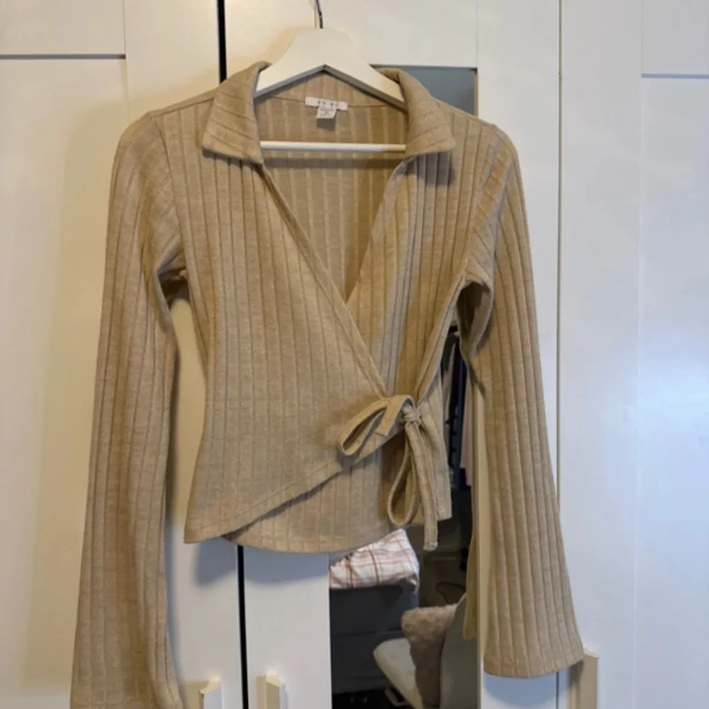 Säljer en superfin beige ribbad omlottopp med knytning i midjan. Den har långa ärmar och en snygg krage som ger en stilren look. Perfekt för både vardag och fest! 🧡. Neuletakit & Villapaidat.