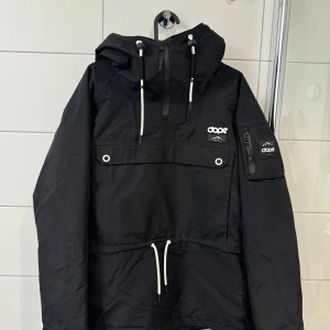 Svart skidjacka från Dope - Säljer en svart jacka från Dope i mycket bra skick. Den har en stor huva med justerbara snören och en praktisk dragkedja framtill. Jackan har en ficka med knappdetaljer och dragkedjor på bröstet och en extra ficka på ärmen. Ficka för skidkort finns på underarmen. Snörning finns i midjan. Och snöskydd. Hör av er vid fler frågor eller bilder. 