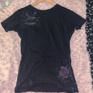 Svart t-shirt med tryck från Sinful - Säljer en svart t-shirt från Sinful i 100% bomull. Jag har knappt använt den så den har bara legat i garderoben ett tag, men köpte den secondhand på auktion så har förmodligen använts en del.  Den har ett coolt tryck med fåglar och rosor i lila och blått, samt texten 'Love & Pride'. Jag är ganska säker på att det är meningen att den ska se lite ”trasig” ut då det är ganska strukturerat runt kragen och nedre delen av tröjan, men bra att veta om man inte skulle gilla det. 