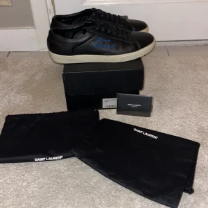 Svarta sneakers från Saint Laurent - Säljer ett par svarta sneakers från Saint Laurent i mycket bra skick. Skorna har en stilren design med Saint Laurent-logga i blått på sidan. De är gjorda i skinn och har en vit sula. Perfekta för både vardag och fest! Kommer med originalförpackning och dustbags.