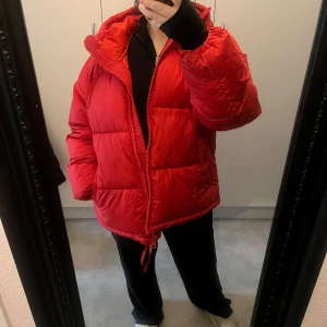 Röd pufferjacka - Säljer en snygg röd pufferjacka i bra skick. Den är perfekt för kalla vinterdagar och har en dragkedja framtill. Jackan är oversized och har en mysig huva. Perfekt för att hålla värmen och se stilren ut samtidigt! Har små fläckar men inget som syns