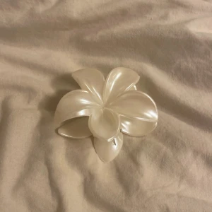Vit hårklämma i blomform - Säljer en superfin vit hårklämma formad som en blomma. Perfekt för att sätta upp håret på ett stilfullt sätt. Klämman är i plast och har en glansig yta som ger en elegant touch. Den är både praktisk och snygg för vardagsbruk eller festliga tillfällen.