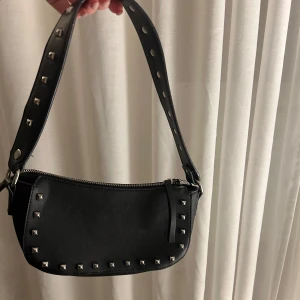 Svart axelväska med nitar - Snygg svart axelväska med coola nitar som detaljer. Från Nelly! Väskan har en dragkedja upptill och en axelrem. Perfekt för en rockig look eller en utekväll. Väskan är i mycket bra skick och redo för nya äventyr!