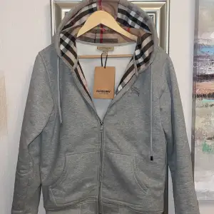 Säljer en snygg grå hoodie från Burberry med det klassiska rutiga mönstret i luvan. Den har dragkedja framtill och är i mycket bra skick. Sparsamt använd och perfekt för höst och vår när du vill vara både stilren och bekväm. Passar till både vardag och lite finare tillfällen. Den är självklart äkta då jag är noga med att sälja och köpa äkta kläder. Burberry lapp kvar som på bilden från när den va ny