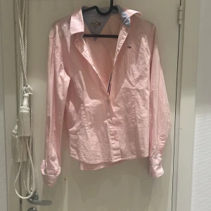 Rosa skjorta från Tommy Hilfiger - Säljer en snygg rosa skjorta från Tommy Hilfiger i bomull. Den har långa ärmar och klassiska knappar framtill. Perfekt för både vardag och fest! Skjortan har en liten logga på bröstet och en stilren design. Passar bra till både jeans och chinos. 🩷