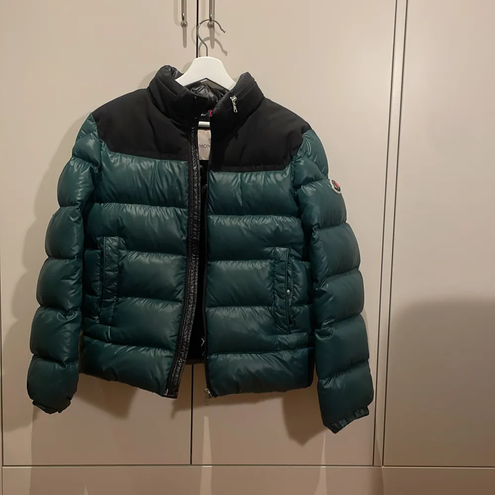 Säljer en snygg grön och svart dunjacka från Moncler. Jackan har en cool pufferstil med dragkedja och Moncler-logga på ärmen. Perfekt för kalla dagar och håller dig varm och stilren. Den är i mycket bra skick och redo för nya äventyr!. Jackor.