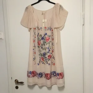 Ivory klänning från Monsoon - Säljer en vacker ivory klänning från Monsoon med blommigt broderi. Den är i helt nytt skick💕Klänningen har kortärmade plisserade ärmar och en lös passform. Perfekt för sommarens festligheter! Orginal pris : 1349kr