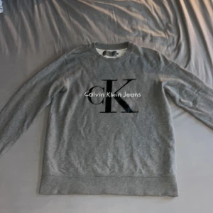 Calvin Klein tjocktröja  - En Calvin Klein Jeans tjocktröja i färgen grå med svart/vit tryck. Storleken är L men passar även bra på M. Tröjan är knappt använd (3-4 gånger max) och har ett minimalt litet hål som är fint lagat (knappt synligt). Nypris 500kr