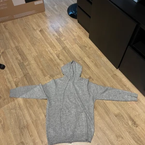 Grå hoodie - Säljer en stilren grå hoodie med dragsko i huvan. Tröjan har långa ärmar och ribbade muddar vid ärmslut och nederkant. Perfekt för en avslappnad look.