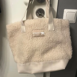 Beige handväska från Kippis - Säljer en mysig beige shoppingväska/handväska från Kippis med ett fluffigt yttre och stilrena handtag. Väskan har ett unikt mönster på insidan och är perfekt för att bära med sig allt man behöver. Den har en praktisk storlek  och är lätt att matcha med olika outfits. bredd: ca36cm längd: ca 26cm