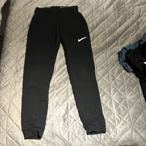Svarta mjukisbyxor från Nike - Säljer ett par svarta mjukisbyxor från Nike med elastisk midja och det klassiska Nike-logotypen i vitt på benet. Perfekta för en avslappnad stil eller träning. Byxorna har en bekväm passform och är gjorda i mjukt material.