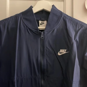 Blå vindjacka från Nike - Snygg blå vindjacka från Nike med dragkedja och vit logga på bröstet. Perfekt för blåsiga dagar och sportiga aktiviteter.