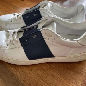 Valentino Open - Snygga Valentino Garavani sneakers i vitt skinn med en bred blå detalj över snörningen. Skorna har en klassisk design med vita snören och en robust sula. Perfekta för en stilren look. 