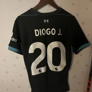 Svart Liverpool fotbollströja - Säljer en svart Liverpool fotbollströja från Nike med 'Diogo J.' och nummer 20 på ryggen. Tröjan har korta ärmar med turkosa detaljer och sponsorer som Standard Chartered och Expedia. Perfekt för fans av LFC!
