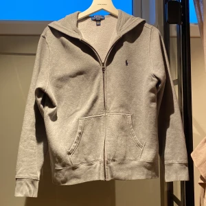 Grå hoodie från Polo Ralph Lauren - Säljer en klassisk grå hoodie från Polo Ralph Lauren med dragkedja och fickor framtill. Den har en broderad logga på bröstet och är perfekt för en avslappnad stil. Passar både för kyliga dagar och som ett lager under jackan. Den är storlek L (14-16)passar 160-168. Det går med lite prutning. Om någon har frågor eller vill köpa hör av er.