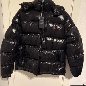 Svart pufferjacka från Ralph Lauren - Snygg svart pufferjacka från Ralph Lauren med glansig finish och ikonisk logga på bröstet. Jackan har en praktisk dragkedja och en mysig huva för extra värme. Perfekt för kyliga dagar. Den är helt oanvänd säljs för att den är för stor på mig.