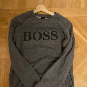 Grå sweatshirt från Hugo Boss - Snygg grå sweatshirt från Hugo Boss med stort BOSS-tryck på framsidan. Tröjan är tillverkad i 100% bomull och har en klassisk rund halsringning. Perfekt för en stilren och bekväm look. Pris kan diskuteras vid snabb affär!