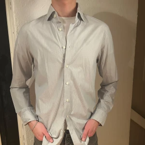Vit och blå randig skjorta - Snygg vit och grå randig skjorta med knappar framtill. Skjortan har en klassisk krage och långa ärmar, perfekt för en stilren look. Passar bra till både jeans och kostymbyxor. Storlek L, 42. 