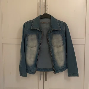 Blå jeansjacka från Contact - Snygg blå jeansjacka med dragkedja och två bröstfickor. Jackan har en klassisk krage och slitna detaljer som ger en cool look. Perfekt för en avslappnad stil.