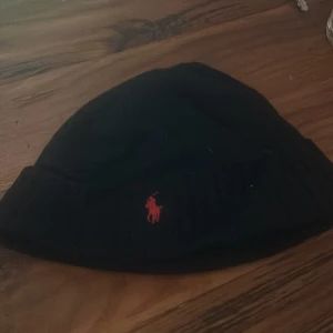 Svart mössa från Polo Ralph Lauren - Snygg svart mössa från Polo Ralph Lauren med röd broderad logga. Tillverkad i mjuk merinoull för extra komfort och värme. Perfekt för kyliga dagar. Skriv vid frågor!! Pris kan diskuteras vid snabb affär