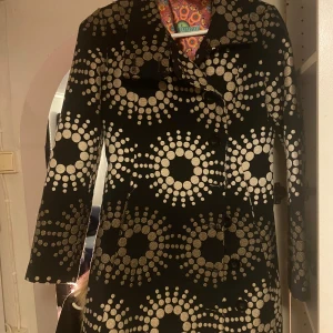 Svart kappa från Desigual med mönster - Snygg svart kappa från Desigual med unikt cirkelmönster i guld. Den har långa ärmar och en färgglad insida som ger en extra touch. Perfekt för att sticka ut i mängden!