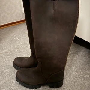 Bruna höga stövlar med dragkedja - Bruna höga stövlar i Mocka med robust sula och dragkedja längs baksidan. De har även ett dekorativt spänne upptill för extra stil. Perfekta för höst- och vinterbruk.