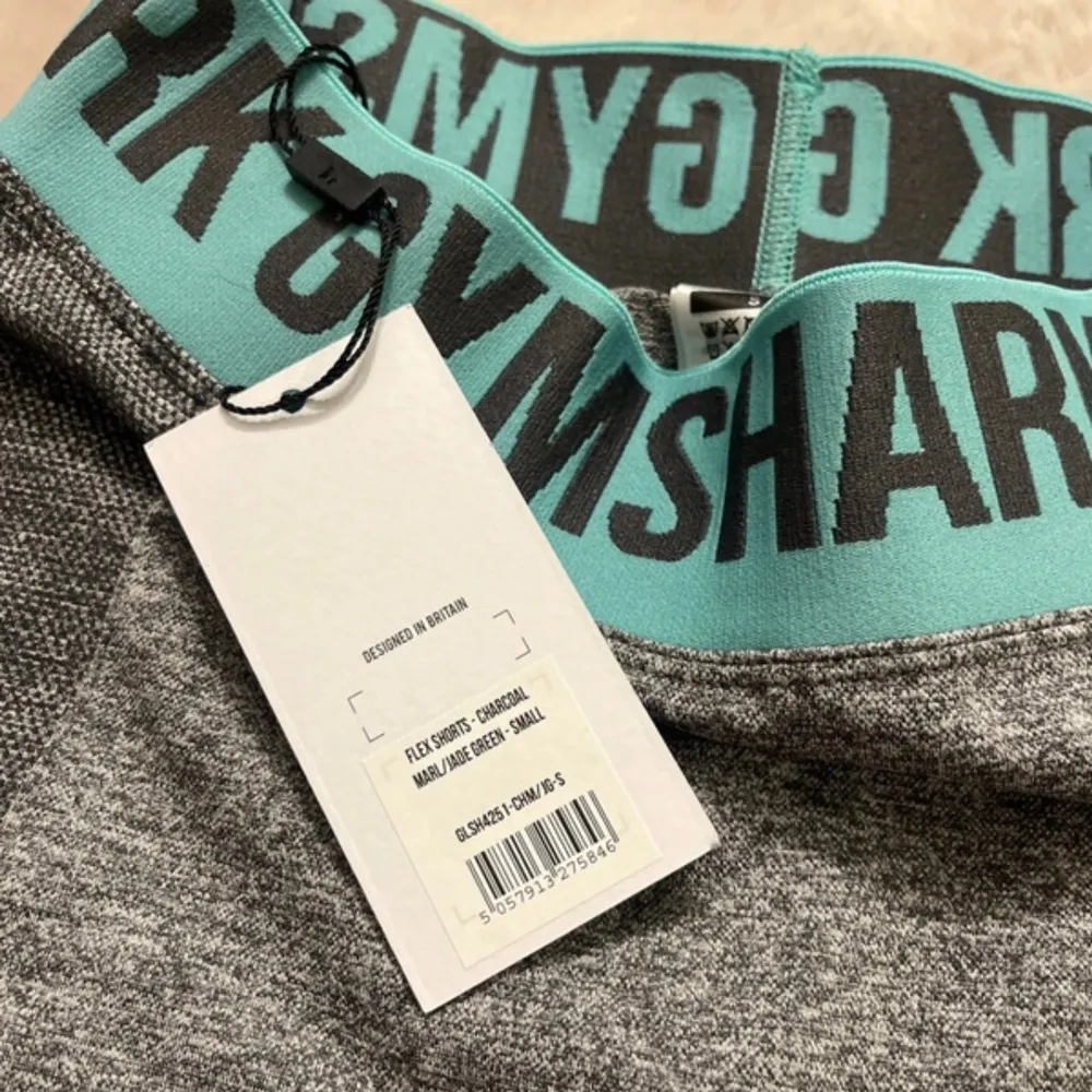 Snygga grå träningsshorts från Gymshark med en bred, turkos resårmidja och svart text. Perfekta för gymmet eller löprundan. De är tillverkade i ett stretchigt material som ger en tight passform. Alldrig använda!. Sportswear.