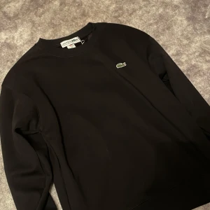 Svart sweatshirt från Lacoste - Säljer en stilren svart sweatshirt från Lacoste med det klassiska gröna krokodilmärket på bröstet. Tröjan har långa ärmar och en rund halsringning, perfekt för en avslappnad look. Storlek M, aldrig använd!!