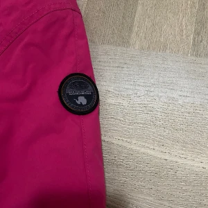 Rosa jacka från Napapijri - Snygg rosa jacka från Napapijri med huva och grått foder. Jackan har en praktisk dragkedja på sidan och en stor ficka framtill med Napapijri-logga och norska flaggan. Perfekt för kyliga dagar med tejpade sömmar för extra skydd.