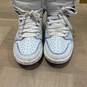 Nike Air Jordans i vitt och blått - Snygga Nike Air Jordans i vitt med blå detaljer. Skorna har en hög modell med snörning och perforerade detaljer på tån. Den blå swooshen och kragen har ett unikt mönster som ger en cool touch. Använd fåtal gånger