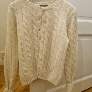 Vit stickad kofta från Brandy Melville - Säljer en mysig vit stickad kofta från Brandy Melville med flätmönster. Koftan har långa ärmar och knappar framtill. Perfekt för kyliga dagar och att matcha med olika outfits.