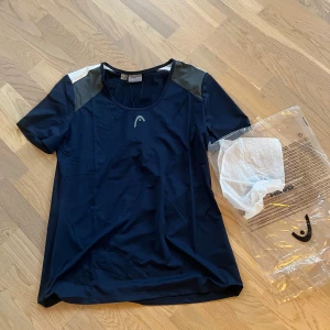 Mörkblå t-shirt från Head - Dam - Säljer en mörkblå t-shirt från Head i storlek XL. Perfekt för träning med korta ärmar och rund hals. Materialet är lätt och andas bra, vilket gör den idealisk för aktiva dagar. Helt ny, nypris ca 450kr