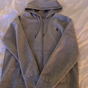 Grå hoodie från Polo Ralph Lauren - Säljer en stilren grå hoodie från Polo Ralph Lauren med dragkedja och justerbar huva. Tröjan har en broderad logotyp på bröstet och praktiska fickor framtill. Köptes på johnells för 2299kr. Storlek S