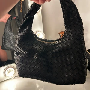 Svart flätad handväska - Hej detta är en svart handväska med flätat mönster och dragkedja. Perfekt för att ge en stilren touch till din outfit. Den är i ett jättebra skick! 