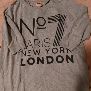 Grå tröja från ONLY - Säljer en grå tröja från ONLY med tryck av städerna Paris, New York och London. Tröjan har medel långa ärmar och en avslappnad passform. Perfekt för en casual look!