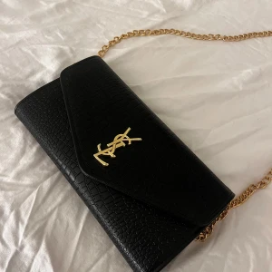 Kuvertväska INTE ÄKTA - Elegant svart kuvertväska med mönster och guldfärgad kedja. Väskan har en stilren design med ett guldemblem på framsidan och en praktisk dragkedja inuti. Perfekt för att ge en lyxig touch till din outfit. INTE äkta. Har några små märken men knappast synliga. Jättefin