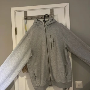 Grå hoodie med dragkedja - Säljer en grå hoodie med dragkedja och huva. Tröjan har långa ärmar och ribbade muddar vid ärmslut och nederkant. Perfekt för en avslappnad stil.