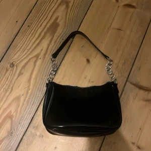 Svart axelväska med kedjedetaljer -  svart axelväska i glansigt material med silverfärgade kedjedetaljer. Väskan har en dragkedja upptill, dragkedjan kan vara svår att stänga OM man tar i stänger man den försiktigt är de aldrig några problem inget slitage 