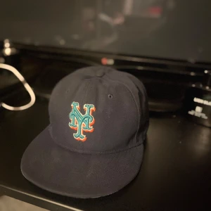 Mörkblå new era keps med broderad logotyp - Snygg mörkblå keps med en broderad logotyp framtill och ett 50-årsjubileumsmärke på sidan. Perfekt för sportiga tillfällen eller som en stilren accessoar.