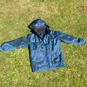 Blå vindjacka från Ski Industries - Snygg blå vindjacka från Ski Industries med huva och kamouflagemönster. Jackan är vindtät och vattenavvisande, perfekt för utomhusaktiviteter. Den har fickor på insidan, ärmarna och praktiska fickor framtill.