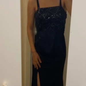 Svart glittrig långklänning - Elegant svart långklänning med glittriga detaljer och smala axelband. Klänningen har en stilren slits framtill som ger en sofistikerad look.