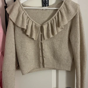 Volang kofta  - Säljer en söt beige kofta med ribbad struktur och volangdetalj runt halsen. Koftan har långa ärmar och knappar framtill. Perfekt för en stilren och mysig look.