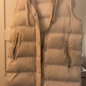 Beige dunväst från Stories - Snygg beige dunväst från Stories i storlek XS. Västen har dragkedja och knappar framtill samt praktiska fickor med dragkedja. Perfekt för kyliga dagar, den är mycke snyggare när man har på sig den.
