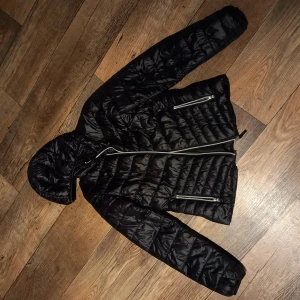 Svart pufferjacka med huva - Snygg svart pufferjacka med huva och dragkedja framtill. Sparsamt använd. 