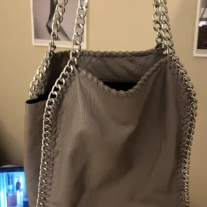 Grå axelväska med kedjedetaljer - stella mccartney liknande Snygg grå axelväska med silverfärgade kedjehandtag och dekorativa sömmar längs kanterna. Perfekt för att ge en stilren touch till din outfit. ‼️pris går att diskutera ‼️