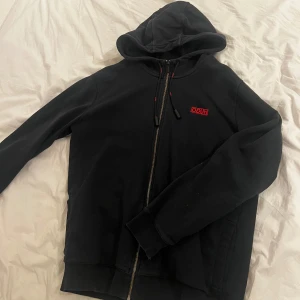 Svart hoodie från Hugo Boss - Snygg svart hoodie från Hugo Boss med röd logga på bröstet. Den har en dragkedja framtill och justerbar huva med snören. Perfekt för en avslappnad stil.